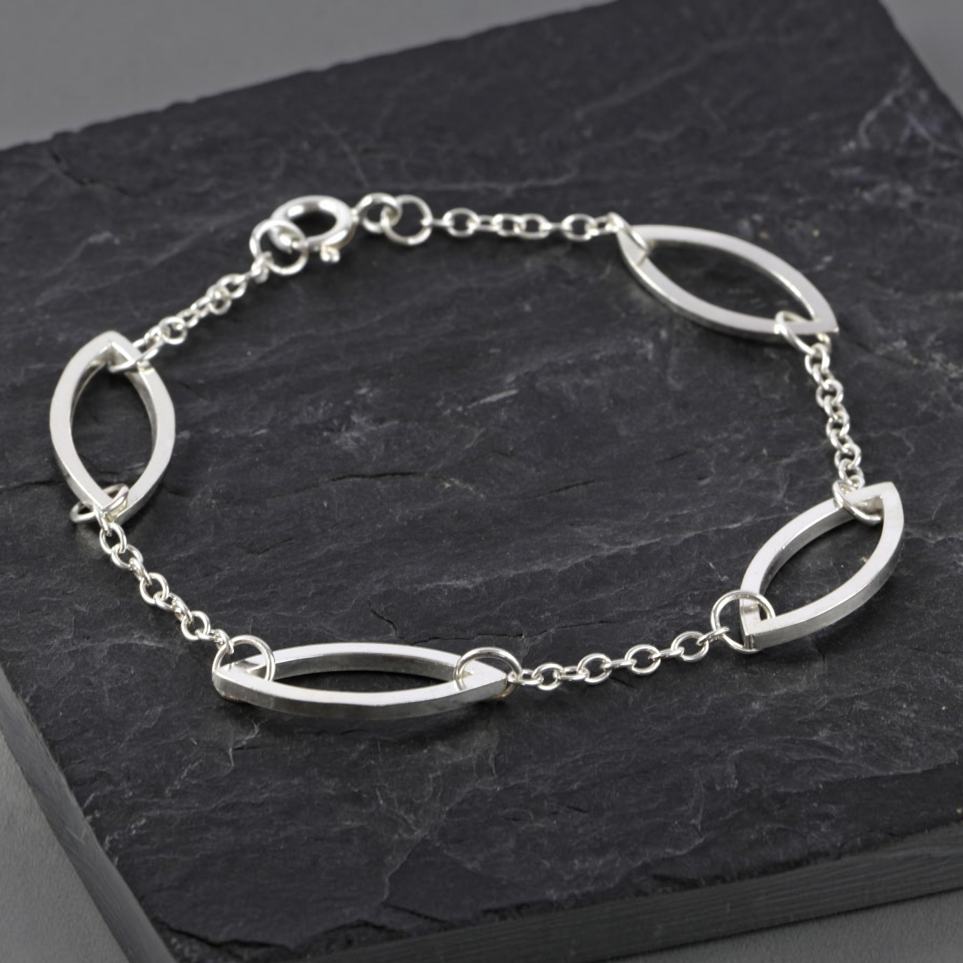 A silver chain bracelet with open marquise links, displayed on a dark textured slate block.