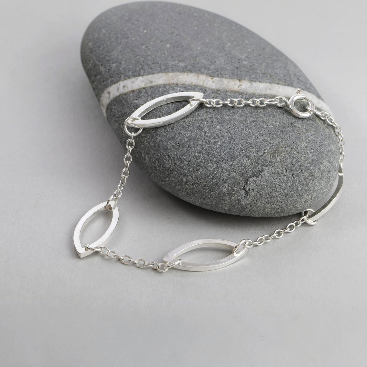 A hand made silver bracelet with marquis shaped links, displayed over a smooth gray stone on a light gray surface.