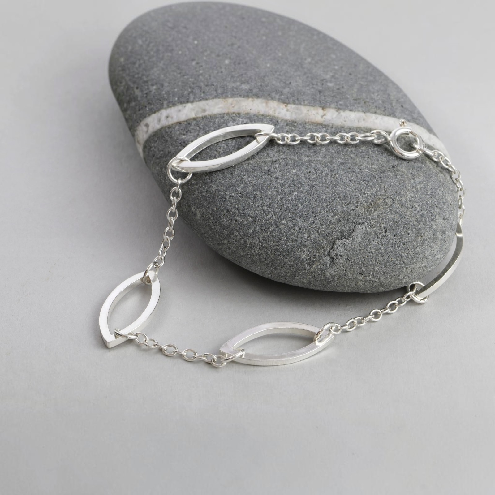A hand made silver bracelet with marquis shaped links, displayed over a smooth gray stone on a light gray surface.