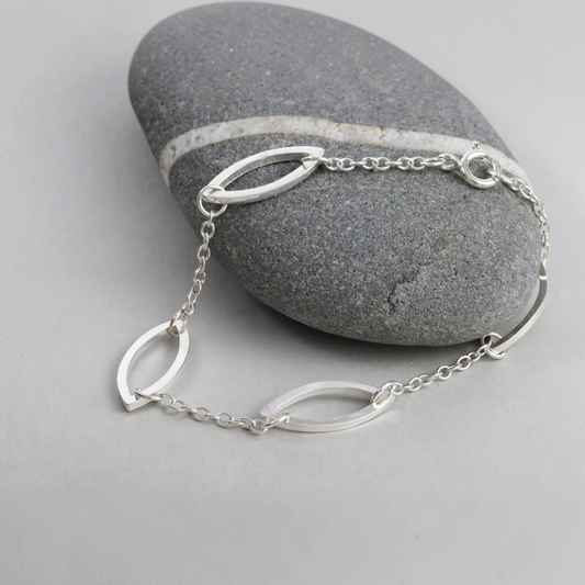 A hand made silver bracelet with marquis shaped links, displayed over a smooth gray stone on a light gray surface.