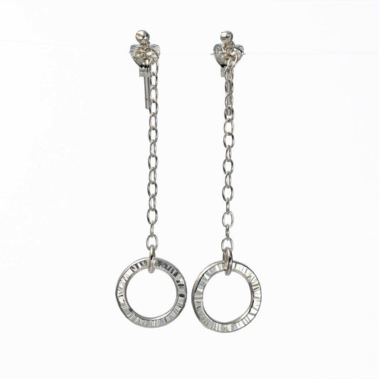 A pair of silver hoop earrings with intricate detailing, suspended on a delicate chain.